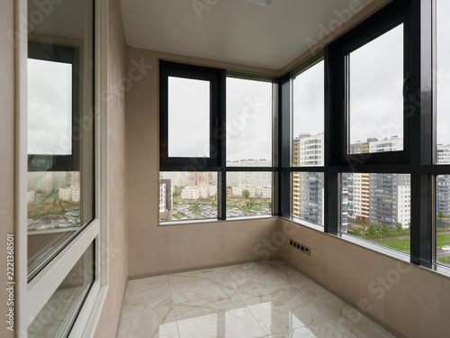 Bright enclosed balcony interior with large glass windows framing urban city skyline, modern high rise architecture, daylight streaming across glossy marble floor and beige walls