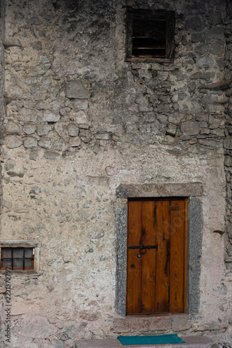Rustic wooden door and small barred window on an ancient stone wall facade in the medieval village of Canale di Tenno, Italy.