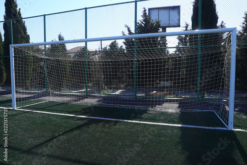 High metal fencing and green trees surround the pitch under a bright blue sky on a sunny day . A white soccer goal with a net stands on green artificial turf at an outdoor sports field.