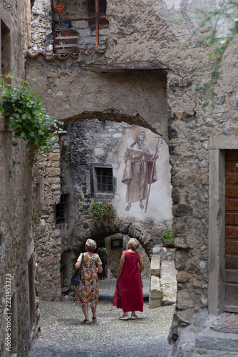Tourists walking through an ancient stone vaulted alleyway with historic wall fresco in the medieval village of Canale di Tenno, Trentino, Italy.