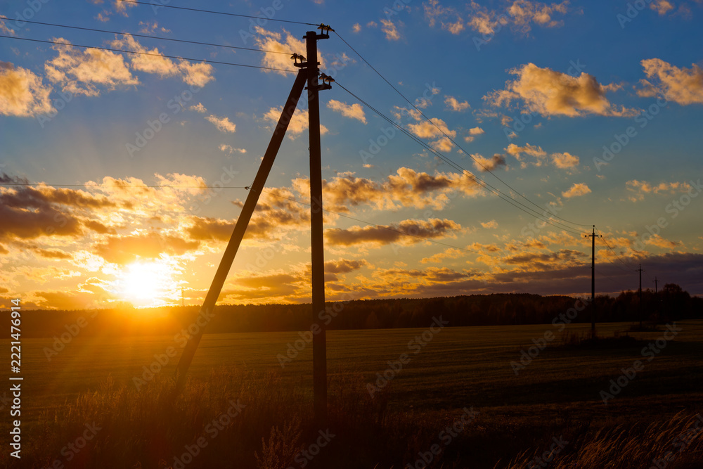 Fototapeta premium Utility poles silhouette against a vibrant October sunset over open autumn fields, highlighting electrical power lines crossing the rural landscape during golden hour