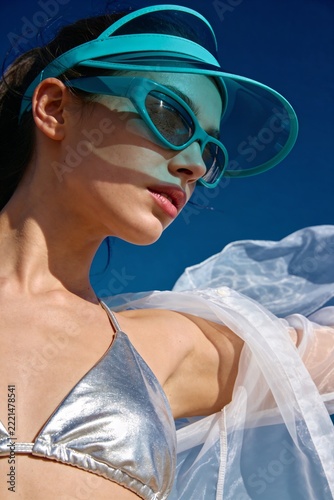 Model poses in silver bikini and sunglasses at beach during sunny day while wind blows through sheer cover-up