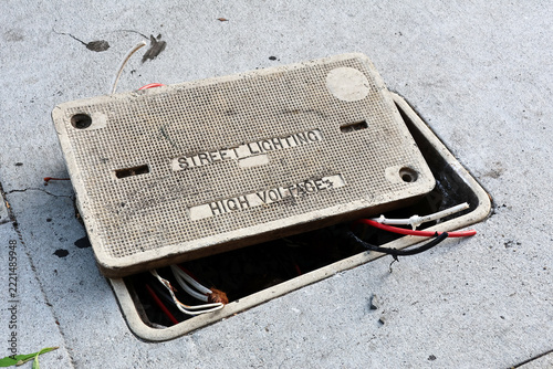 Open electrical utility vault with exposed cables beneath a high voltage street lighting warning on the sidewalk