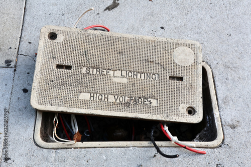 Open electrical utility vault with exposed cables beneath a high voltage street lighting warning on the sidewalk