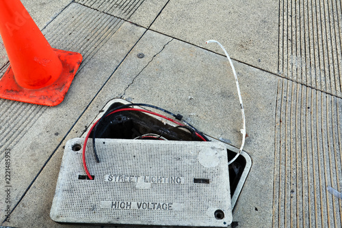 Open electrical utility vault with exposed cables beneath a high voltage street lighting warning on the sidewalk