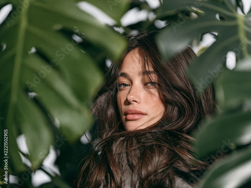 A freckled young woman with wavy brown hair in burnt‑orange garment gazes at viewer, framed by lush large green tropical plant leaves in soft natural light.