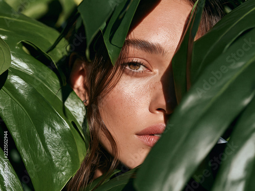 A freckled young woman with wavy brown hair in burnt‑orange garment gazes at viewer, framed by lush large green tropical plant leaves in soft natural light.
