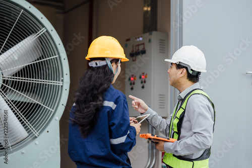 Two industrial workers in hard hats discuss maintenance by a large ventilation unit and control panel inside a plant, using a clipboard and safety gear during an inspection.
