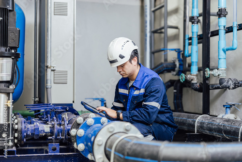 Engineer in protective gear using a tablet to inspect industrial piping and valves in a plant room.