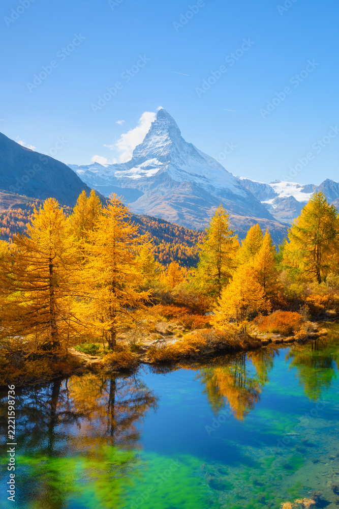 Fototapeta premium Vertical photo. Matterhorn mountain in Switzerland reflects in a calm alpine lake. Scenic mountain landscape with colorful autumn trees under a clear sky in daytime.