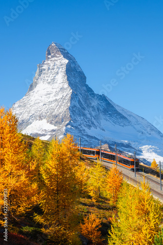 Vertical photo. Passenger train on a railway in Zermatt, Switzerland. Autumn nature in the Alps. Train passes through autumn forest with Matterhorn peak in background.
