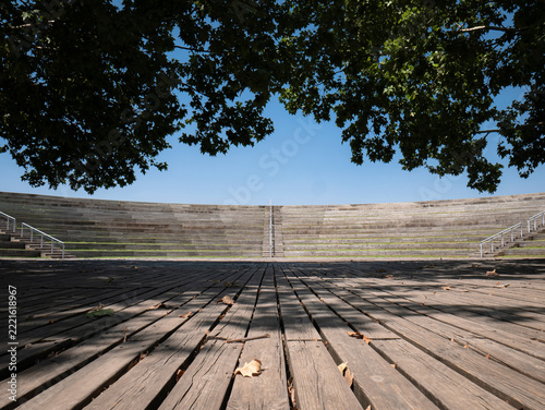 View of the open-air amphitheater in Madrid's Manzanares Linear Park, featuring curved tiered seating with wooden benches, staircases with railings, and a spacious stage with a wooden floor