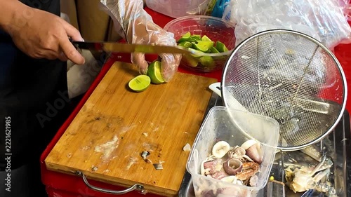 Close up shot of street food vendor hand in glove putting fresh sliced green lime pieces from a wooden board into a plastic bowl at a market stall.
