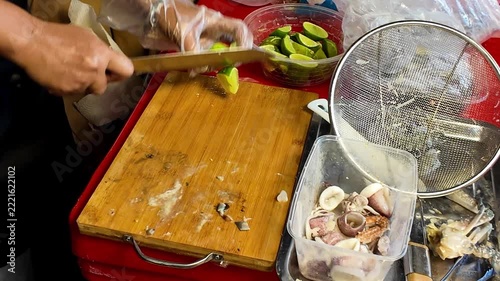 Close up shot of street food vendor hands using a sharp knife to cut fresh green lime fruits into pieces on a wooden board at a market stall.
