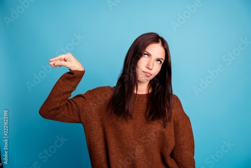 Young woman in brown sweater rolling her eyes and making a dismissive hand gesture against a blue background expressing sarcasm, boredom, annoyance and playful frustration