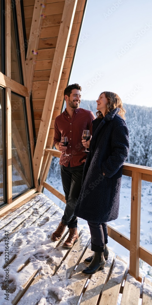 Fototapeta premium Casual winter balcony portrait of smiling couple with wine glasses at a mountain lodge for romantic getaway background and travel design