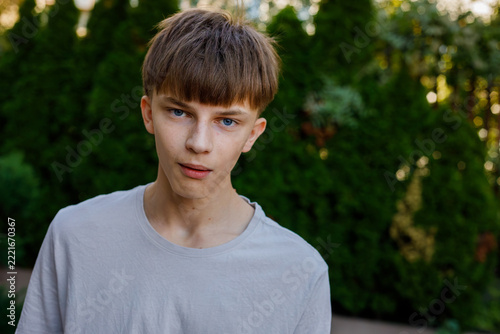 Serious teenage boy with brown hair and bangs in gray shirt looking at camera, outdoor portrait against green trees, calm neutral expression of a boy, grainy film texture, high quality photo