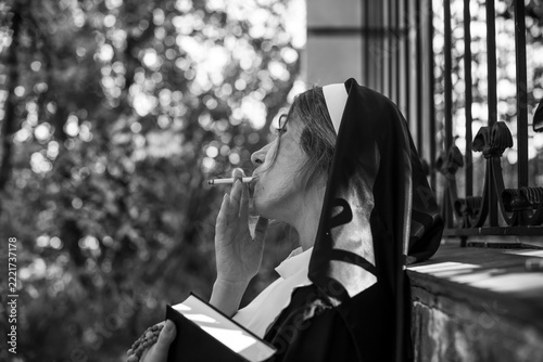 Thoughtful woman wearing a traditional nun inspired habit and holding a cigarette. Dramatic conceptual portrait exploring faith, personal identity, inner conflict, rebellion and the contrast 