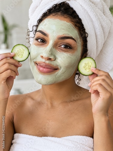 Smiling woman wearing green facial mask and holding fresh cucumber slices