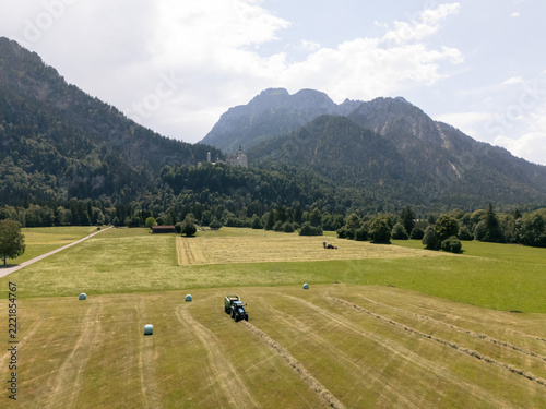 Aerial view of a tractor working in a hay field with hay bales, surrounded by lush green meadows and the dramatic peaks of the Tiroler Zugspitz Arena in Tirol, Austria.