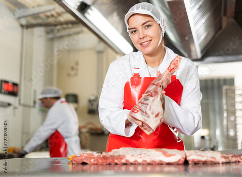 Obraz na plátně Happy young female butchery worker in white uniform and red apron standing at cu