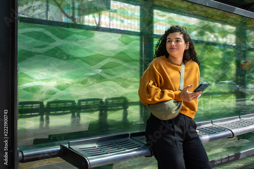Smiling woman tourist wearing a fanny pack waits at a bus stop, holding a phone and looking into the distance; a young traveler using public transport to explore the city