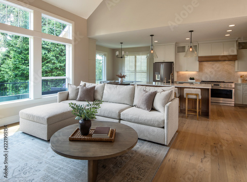 An open-plan living room featuring a beige sectional couch with a chaise lounge, a round wood coffee table, large windows overlooking lush trees, and high vaulted ceilings.
