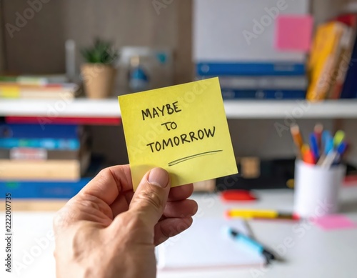 Hand Holding Sticky Note with Maybe to Tomorrow Text on a Bright Office Desk Setting