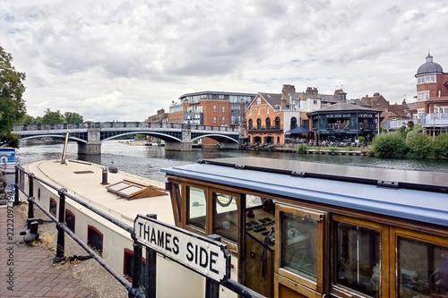 Windsor Bridge and traditional narrowboat moored along the scenic River Thames in Windsor, Berkshire, England, featuring historic architecture and a cloudy sky.