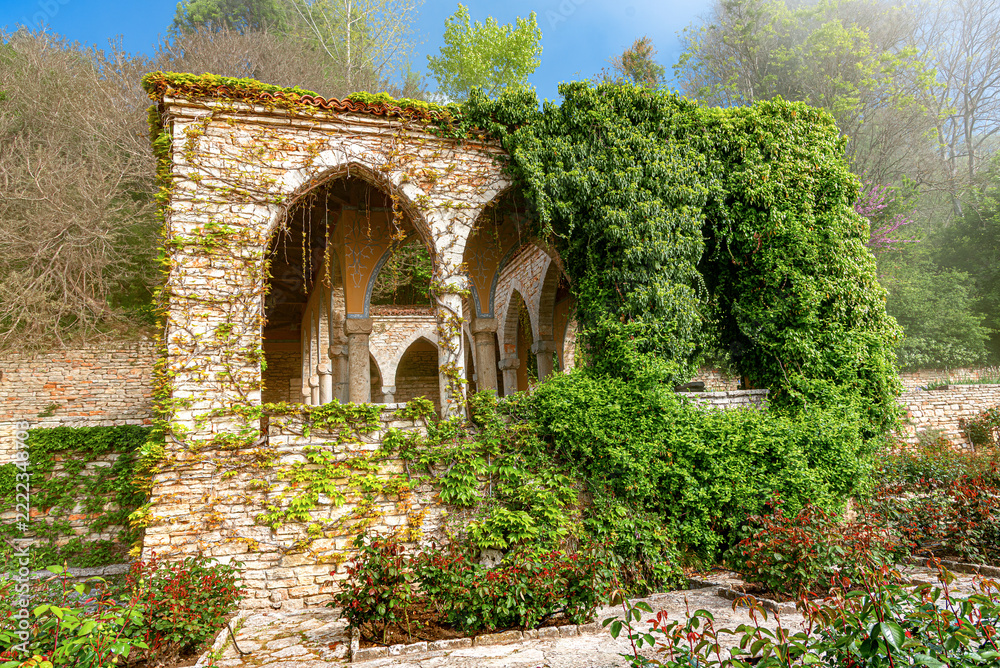 Fototapeta premium An ancient stone pavilion with arched openings covered in lush green ivy stands in a sunny garden.