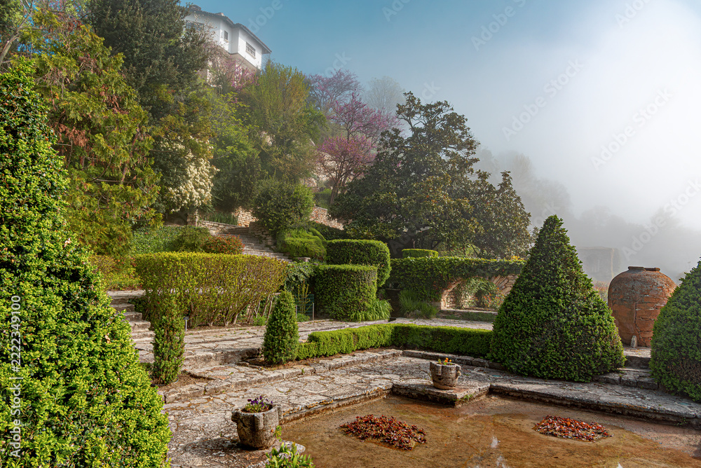 Naklejka premium A beautiful manicured garden features green topiary bushes, stone pathways, and a foggy background on a sunny morning.