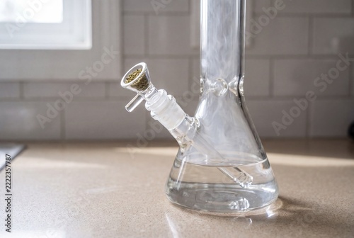 Close-up of a clear glass water pipe filled with water and cannabis, featuring swirling smoke, on a light kitchen countertop with a blurred tiled background and window light.