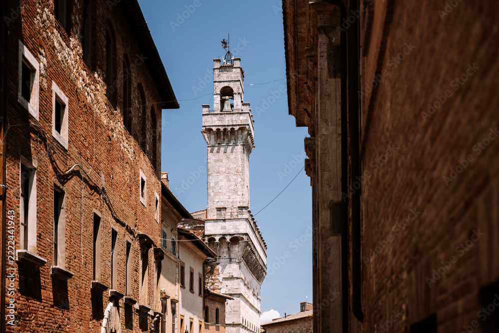 Naklejka premium Historic town hall tower of Palazzo Comunale in Montepulciano, Tuscany, Italy, viewed through a narrow street lined with old brick buildings in the medieval town center.