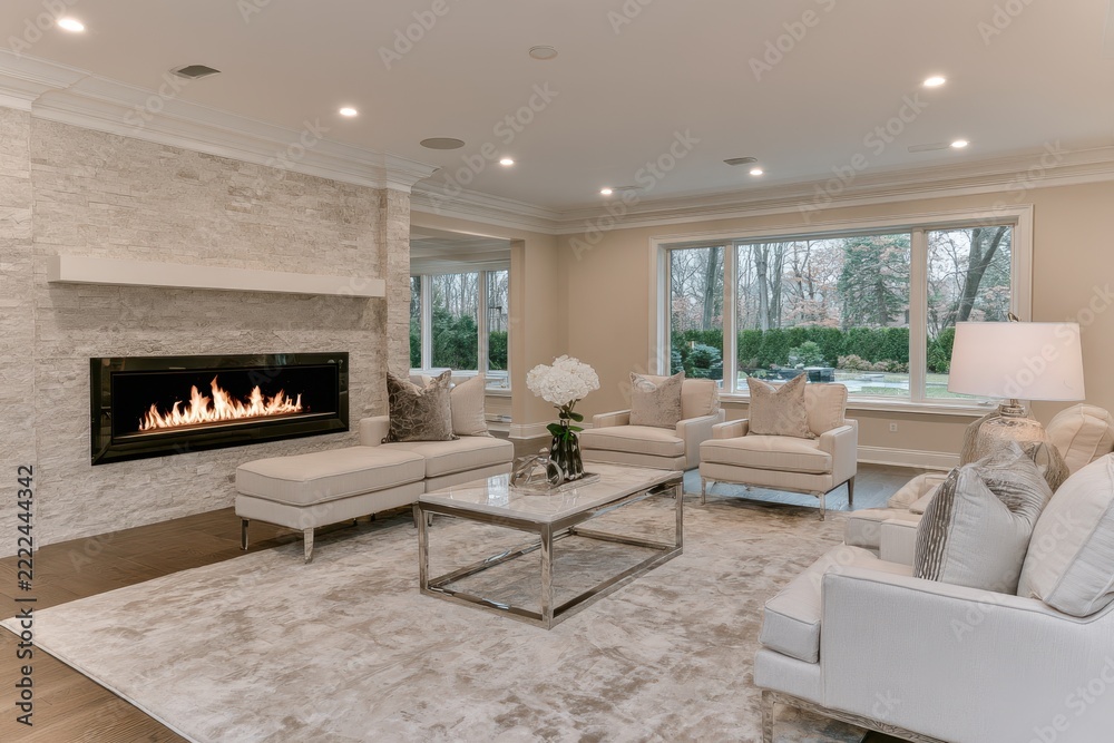 Naklejka premium Elegant neutral living room with modern fireplace, sectional seating, glass coffee table, large windows, and cream furnishings in a refined residential interior