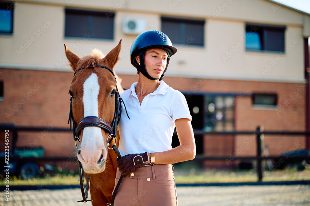 Naklejka premium Woman rider wearing a helmet and riding gear standing with her chestnut horse, fostering a strong bond at an equestrian stable
