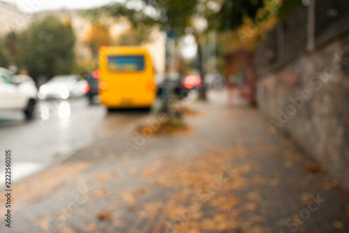 Blurred autumn city street with yellow bus and fallen leaves on wet sidewalk