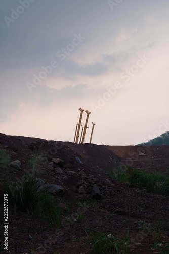 Three yellow piling rigs behind a massive earth mound at a construction site during twilight.