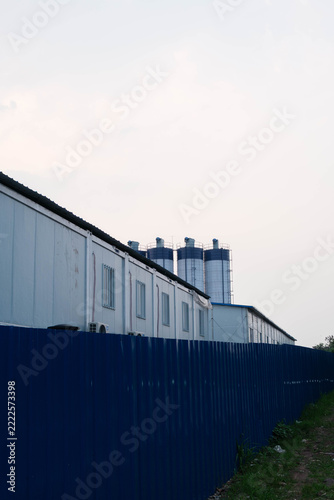 Vertical view of a prefabricated site office behind a blue metal fence with cement silos in the background