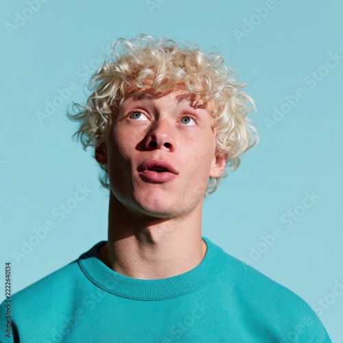 Surprised young man with curly blonde hair in teal sweatshirt on blue background for expressive lifestyle poster and advertising design