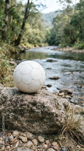White soccer ball resting on a large rock by a flowing forest river, outdoor sports recreation concept.