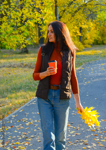 A cheerful woman in a brown vest and a terracotta turtleneck enjoys a walk in the park during the golden autumn; she is holding a bright orange coffee cup and a bouquet of colorful fallen leaves.