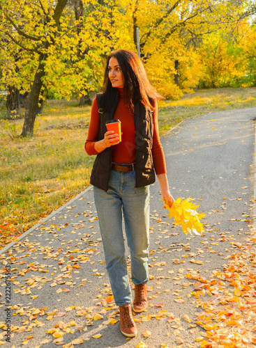 A cheerful woman in a brown vest and a terracotta turtleneck enjoys a walk in the park during the golden autumn; she is holding a bright orange coffee cup and a bouquet of colorful fallen leaves.
