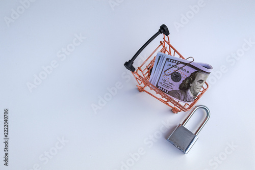 Miniature shopping cart filled with US dollar banknotes beside padlock on white background representing safe shopping and budget lock