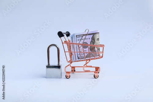 Miniature supermarket shopping cart filled with US dollar banknotes next to locked padlock on white background with copy space