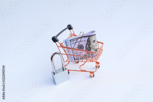 Miniature shopping cart filled with US dollar banknotes beside padlock on white background representing safe shopping and budget lock
