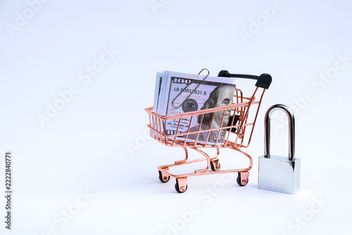 Miniature supermarket shopping cart filled with US dollar banknotes next to locked padlock on white background with copy space