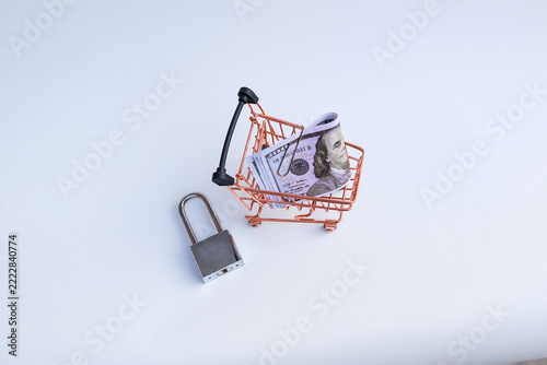 Miniature shopping cart filled with US dollar banknotes beside padlock on white background representing safe shopping and budget lock