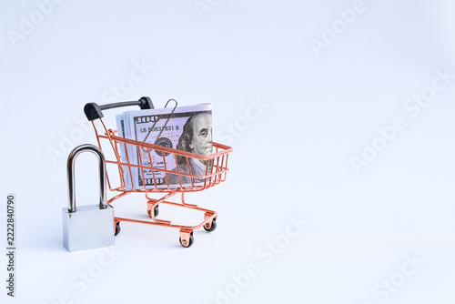 Miniature supermarket shopping cart filled with US dollar banknotes next to locked padlock on white background with copy space