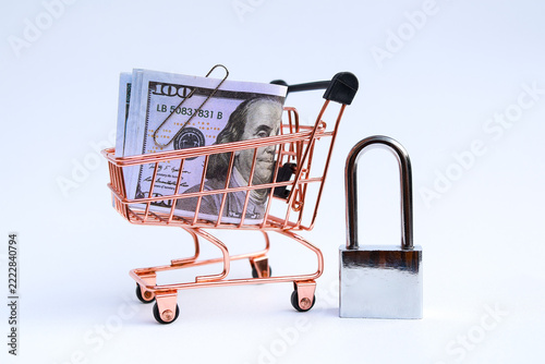 Miniature supermarket shopping cart filled with US dollar banknotes next to locked padlock on white background with copy space