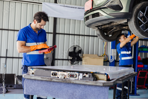 Auto technicians service an electric car in a garage, inspecting a removed EV battery pack on a workbench while using a tablet device, with the vehicle lifted on a hydraulic hoist.